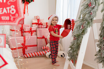 Little girl in Santa Claus hat and red pajamas on the background of Christmas gifts. Waiting for Christmas gifts. Merry Christmas .