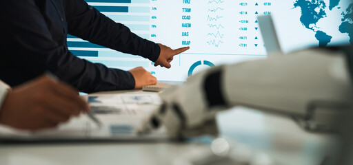 Professionals engage in data analysis, utilizing a robotic hand and digital screen to navigate financial trends and insights during a collaborative workplace discussion.Noogenesis