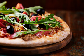 Pizza with salami, tomatoes and mozzarella cheese, black olives and arugula. Rustic wooden background. Soft focus. Copy space.	