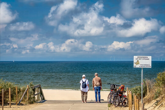 Two people stand at the end of a beach path, looking towards the ocean. Bicycles are parked nearby, and the sky is dotted with clouds.