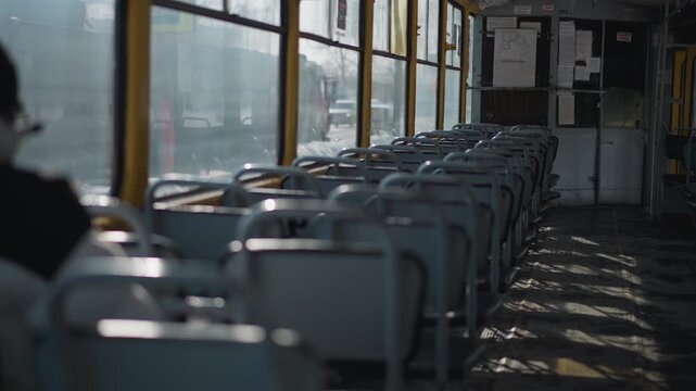 nearly empty train interior shows rows of blue seats in winter light while single passenger rides at back, city cars glide past windows, long shadows stripe worn floor, calm transit mood