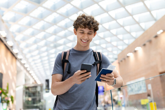 Young Caucasian man with backpack holding passport and smartphone, smiling inside airport terminal. Travel concept of departure, boarding, youth lifestyle and digital routine