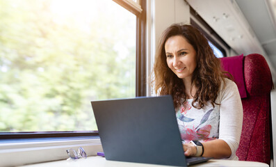 Young Caucasian woman smiling while working on laptop during train travel. Concept of digital lifestyle, remote work, and productive commuting on the go