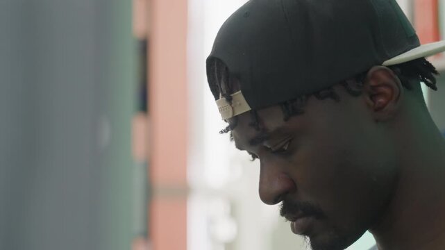 Close up side profile of college student in backward cap by library shelves, sorting book with focused expression, soft blur background and muted colors capturing quiet study mood