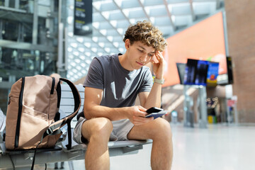Young man with backpack sitting at airport looking worried at passport and smartphone. Represents...
