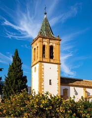 Fototapeta premium A tall, ornate church tower, distinguished by its tan and white stonework, rises against a vibrant blue sky dotted with wispy clouds. Its tiled green roof provides a striking contrast.