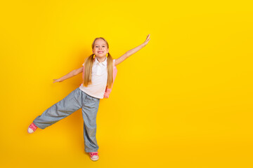 Cheerful Schoolgirl in Casual Clothes Raising Her Arms Playfully Against a Bright Yellow Background