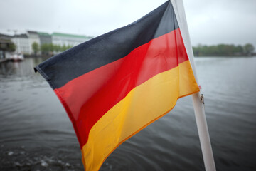 German national flag waving on a white pole by a waterfront. Background includes trees, buildings, and cloudy sky, suggesting an urban setting.