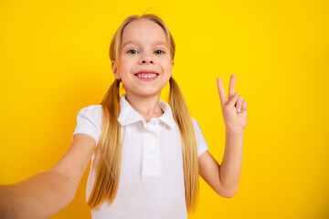 Cheerful schoolgirl showing peace sign gesture in white shirt against yellow background