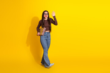 Beautiful young woman holding a patterned book, waving cheerfully, and posing in casual attire against a bright yellow backdrop