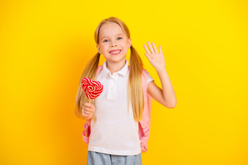 Happy schoolgirl holding a red heart-shaped lollipop and waving against a bright yellow background