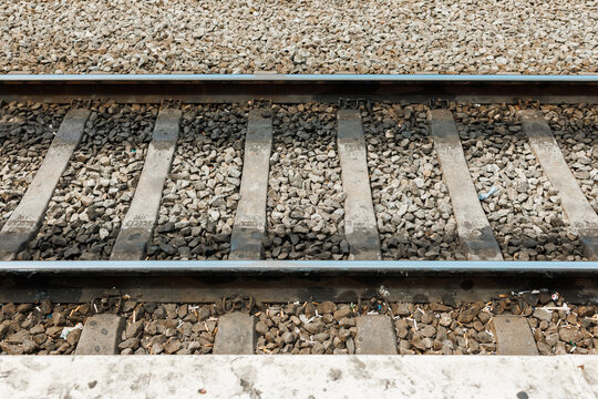 Close-up view of parallel steel railroad tracks with weathered wooden ties embedded in gray gravel, extending from a concrete platform edge. Stock photo