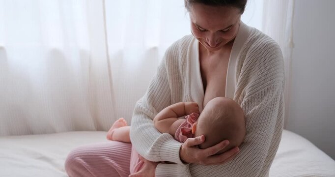 Slow-motion medium shot of a mother breastfeeding her infant daughter while sitting on the bed.