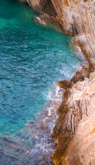 Turquoise seawater laps against rugged layered rock formations on a steep coastline, showcasing striking geological textures and the dynamic meeting of land and sea.