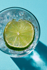Refreshing iced sparkling water with ice cubes and slice of lime top view against pastel blue background. Non-alcoholic drink alternative.