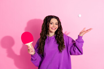 Young woman in purple sweatshirt holding a pink ping pong paddle and showing a playful smile against pink background