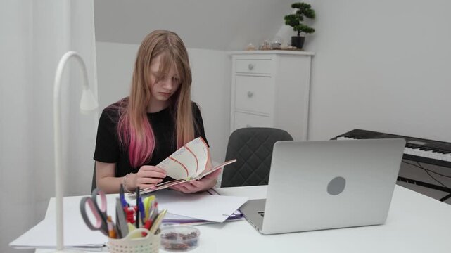Young woman examines her class schedule in a diary while seated at a desk, surrounded by stationery and a laptop, showcasing focused study habits
