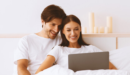 Happy cute european millennial man and woman hugging, sitting on bed, use laptop in white bedroom interior. Couple watch movie together at free time, video call, surf in internet and social networks