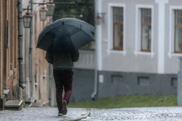 A person walks alone down a quiet cobblestone street in the rain, holding a large black umbrella, surrounded by old buildings and a soft, gray atmosphere.