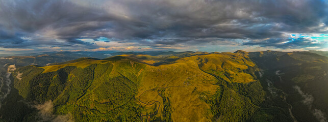 Rolling green hills and forested mountains in Europe under moody sky with dramatic clouds and patches of sunlight aerial panoramic view