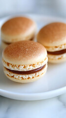 A trio of delicate cream and brown macarons filled with a smooth filling, presented on a crisp white plate, exudes a sense of classic confection perfection.