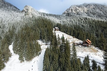 Hikers ascend a snowy path toward a mountain refuge nestled among frosted trees in Romania. Cabins sit on hillsides, surrounded by snow-covered forest and dramatic rock formations