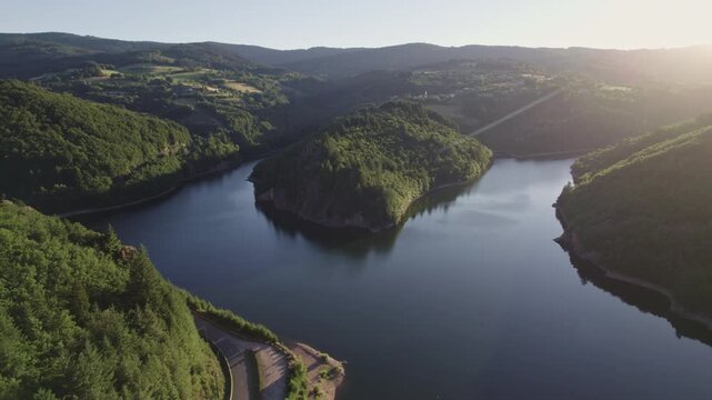 Drone footage of a lake cutting through a mountain, surrounded by pine forests in the French Alps at sunset. Golden light reflects on the water and rugged alpine terrain.