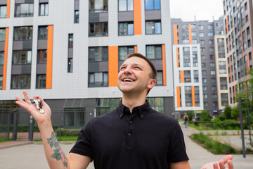 A man joyfully raises his keys, standing in front of a contemporary apartment building with bright orange accents. He expresses excitement about moving into a new home.