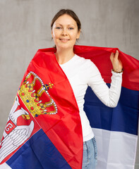Positive patriotic middle-aged woman in casual wear holding state flag of Serbia against gray wall...