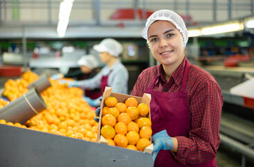 Smiling young female worker in fruit sorting and packing warehouse holding small wooden box of sorted ripe mandarins
