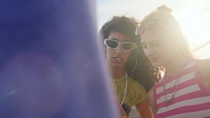 Medium close up slowmo of stylish gen Z diverse girlfriends chatting while looking at smartphone screen, relaxing and enjoying beautiful summer day in city together - Powered by Adobe