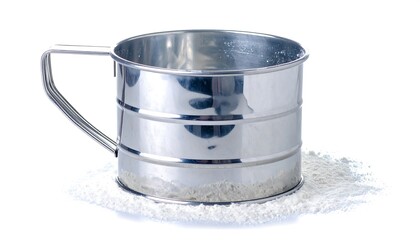 A metallic flour sifter sits atop a bed of fine flour against a white background.