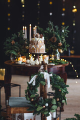 Christmas elegant composition with handmade wreath on wooden chain and beautiful homemade gingerbread house, candles on the table. Family party festive decoration, cozy atmosphere, aesthetics