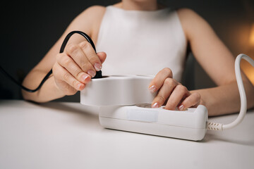 Fototapeta premium Close-up cropped shot of woman plugging black cable into white power strip on white table, with blurred background and soft lighting, creating sense of carefulness and precision.