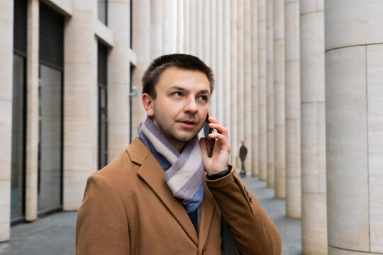 Man dressed in stylish coat with scarf talking on phone while walking through modern city architecture in business setting. businessman, manager, lawyer portrait. adult male 36 years old