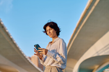 Businesswoman using a mobile phone app while sitting under a highway overpass in the city, enjoying the convenience of technology