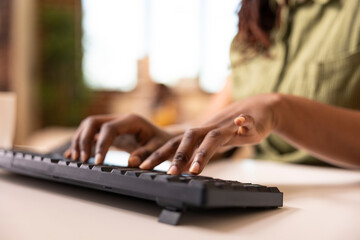 Selective focus on fingers of african american woman inputting business data into desktop pc. Remote work setup in cozy living room with brick wall background showing modern lifestyle.