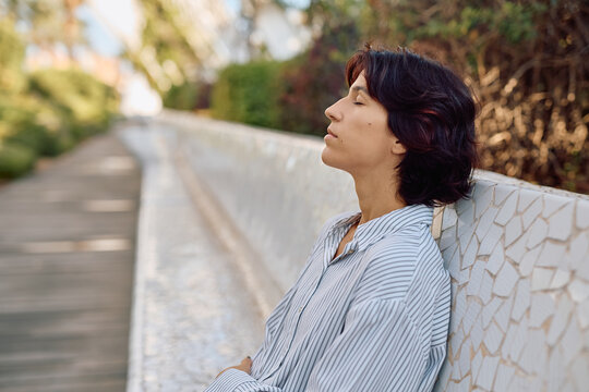 Businesswoman taking a break from work, sitting on a bench with closed eyes, breathing fresh air and enjoying the peace of nature