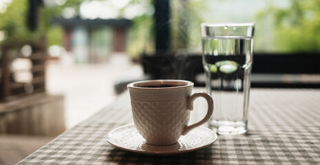 A steaming cup of coffee and a glass of water on a checkered tablecloth in a cozy setting. Strong Balkan coffee in outdoor cafe