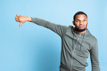 Naklejka premium Displeased irritated person gives a thumbs down sign in studio, showing his negativity and bad vibes against blue background. Young african american guy shows dislike and rejection.