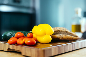 Fresh vegetables including yellow bell pepper, cherry tomatoes, and cucumber arranged on a wooden cutting board with bread and olive oil in a bright kitchen setting