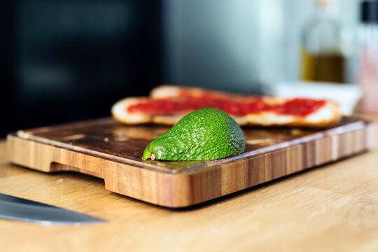 Fresh green avocado resting on a wooden cutting board with a pizza base topped with tomato sauce in the background, showcasing vibrant culinary ingredients and preparation - Powered by Adobe