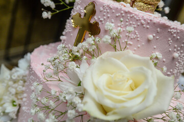 Pink two-tiered round cake with white flowers decorated, gold bear on top and large orthodox gold cross in front.