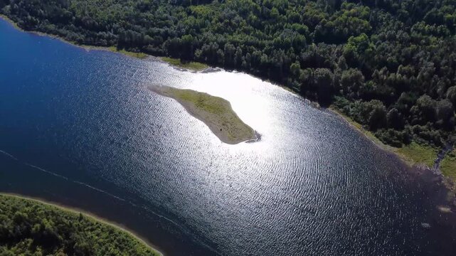 A sandbar forms an island with modest vegetation bathing in the center of the sun's reflection on the surface of a forest lake crossed by small, shimmering waves. Lake Mitis, Quebec, Canada, 2025.