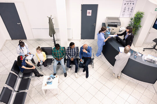 Top view of nurse in blue scrubs and elderly doctor speaking with female hospital receptionist, updating medical records. Clinical lobby showing multiethnic patients preparing for health consultations - Powered by Adobe