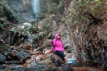 A female hiker in a bright pink jacket resting on rocks near a flowing stream and waterfall. Concept of outdoor adventure, hiking, travel, freedom, and nature exploration in the wilderness.
