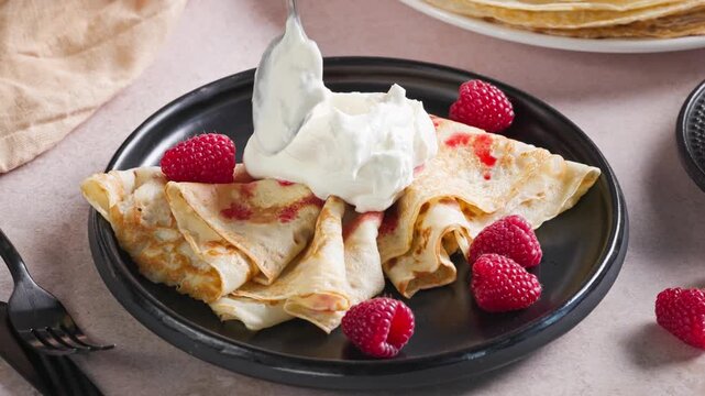 plate of freshly baked crepes decorated with raspberries. cook put whipped yogurt cream on pancakes with spoon for making delicious breakfast, sweet dessert closeup in kitchen