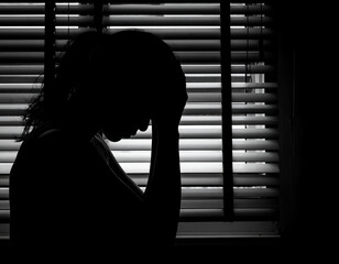 Solemn silhouette of a person overwhelmed by despair, head in hands by window blinds. Evokes deep sadness, anxiety, loneliness, mental health struggles. Black and white.