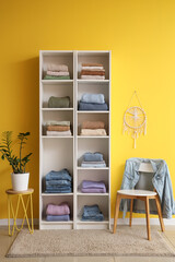 Shelving unit with folded clothes, chair and houseplant near color wall