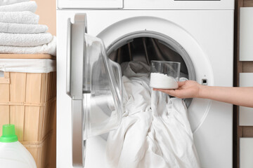 Female hand pouring laundry detergent into washing machine with bed sheets in room